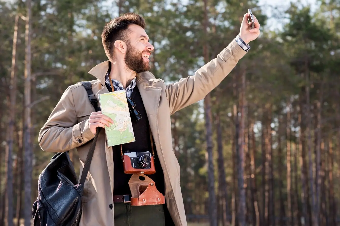 Ein lächelnder junger Mann hält eine Karte in der Hand und macht im Wald mit einem Handy ein Selfie.